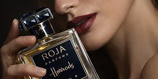 Close-up of a woman with dark red lipstick holding a luxury Roja Parfums Harrods bottle near her face in moody lighting.
