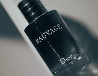 A tilted, close-up shot of a black Dior Sauvage perfume bottle reflecting soft light against a clean, minimalist background.