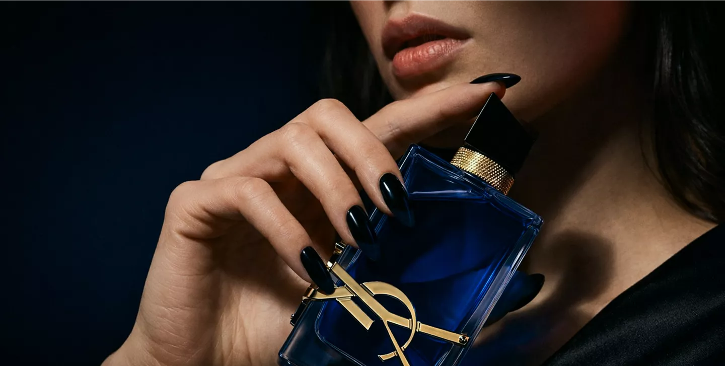 Close-up of a woman's hand with black pointed nails holding a blue YSL perfume bottle against a dark, moody background.