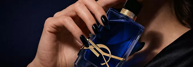 Close-up of a woman's hand with black pointed nails holding a blue YSL perfume bottle against a dark, moody background.