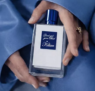 Hands holding Kilian Good Girl Gone Bad perfume bottle with blue cap against soft blue silk fabric background