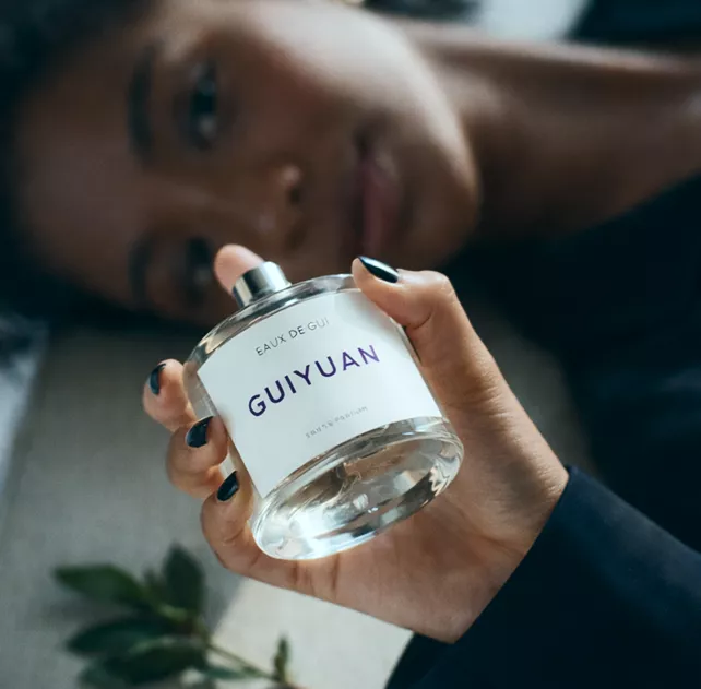 Woman lying down holding Guiyuan Eaux de Gui strong parfum bottle with black nails and a soft, relaxed expression