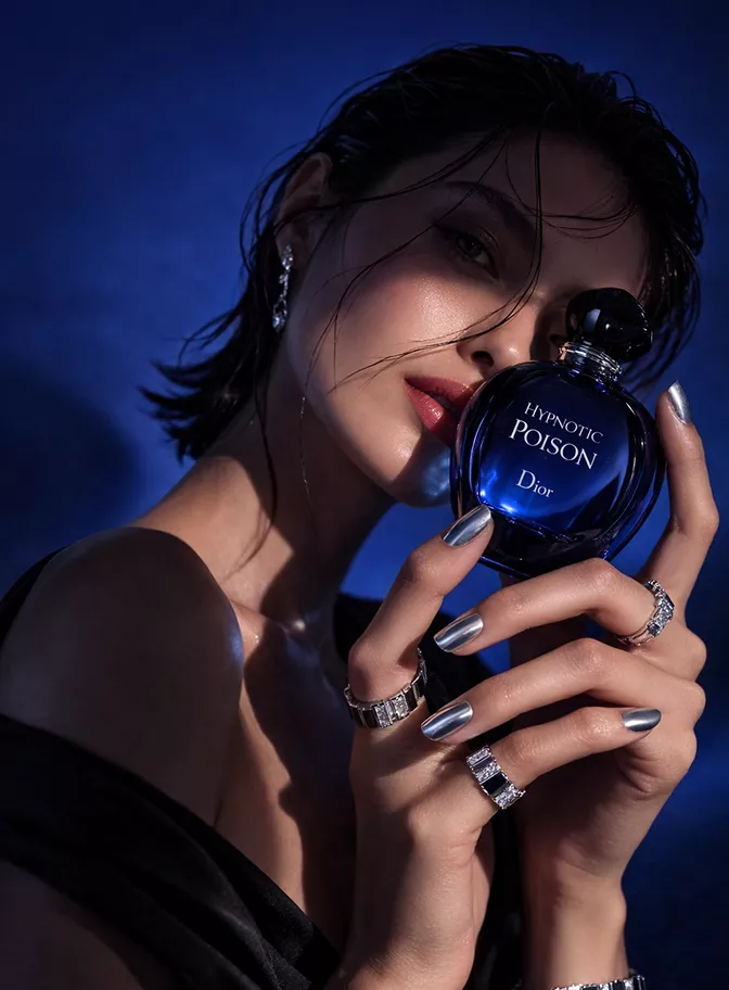 Woman in blue light holding a Dior Hypnotic Poison bottle near her face, featuring silver nails and statement rings.