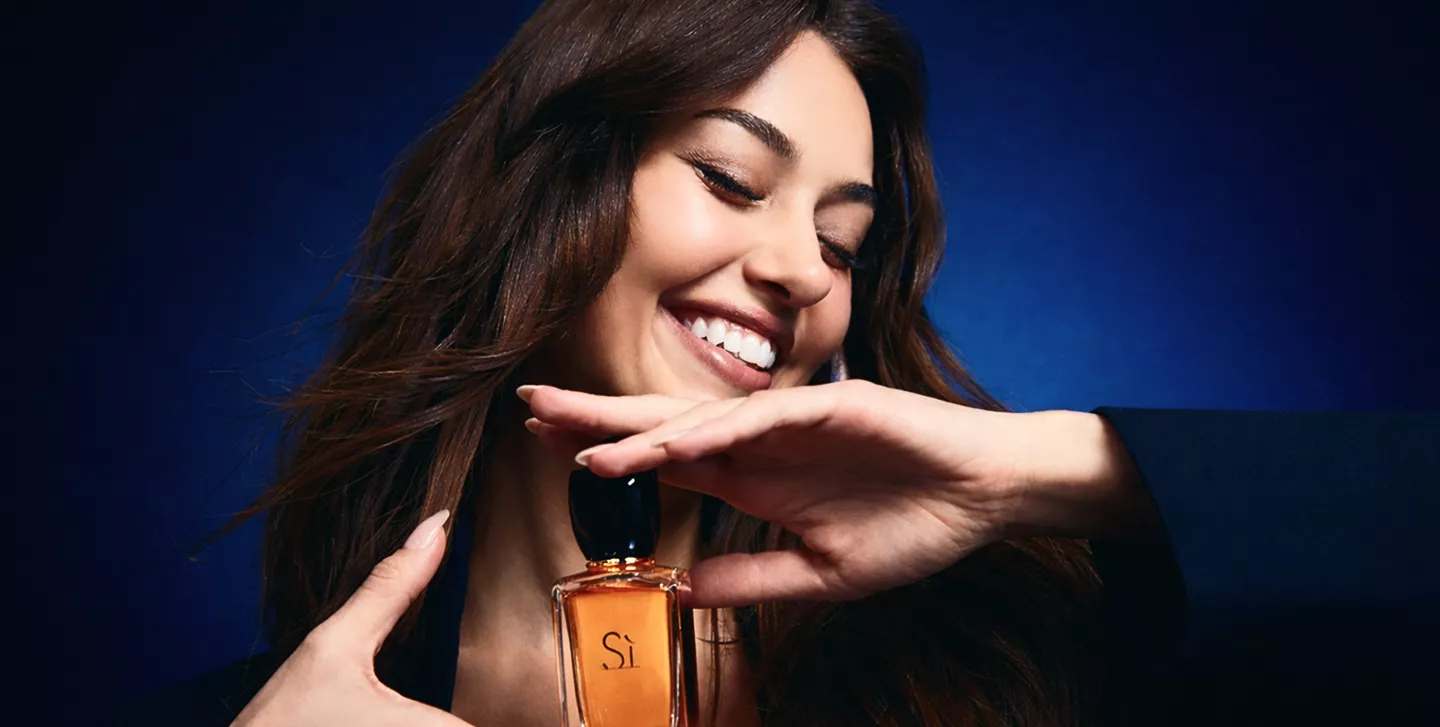 A smiling woman with eyes closed holding an amber Giorgio Armani Sì perfume bottle against a deep blue background.
