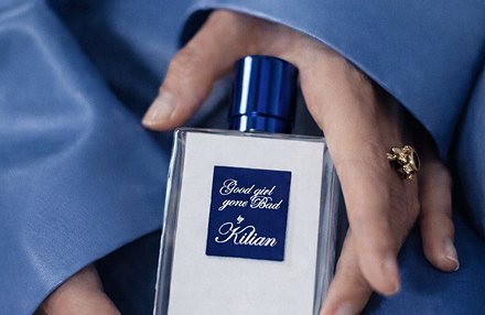 Hands holding Kilian Good Girl Gone Bad perfume bottle with blue cap against soft blue silk fabric background