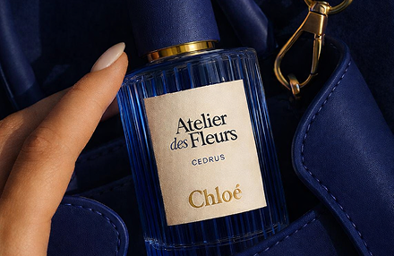 A hand placing a blue ribbed Chlo&eacute; Atelier des Fleurs Cedrus perfume bottle into a matching navy blue leather handbag.